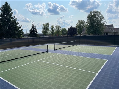 Outdoor sports court with a green and blue surface, white lines, and a tennis net, surrounded by trees and a black fence.