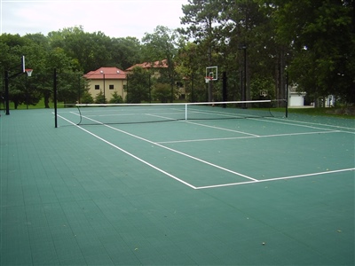 Outdoor residential sports court with a green surface, featuring tennis nets and a basketball hoop.