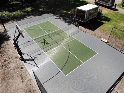 Outdoor sports court with a gray and green surface, featuring a basketball hoop and a tennis net.