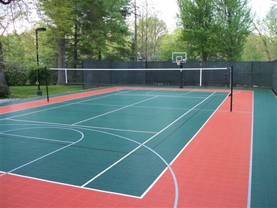 Outdoor sports court with a green and red surface, featuring basketball hoops and a volleyball net, surrounded by trees and greenery.