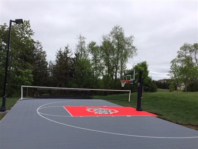 Outdoor sports court with a gray surface and a red center area with a central logo, featuring a basketball hoop and a volleyball net in Watertown.