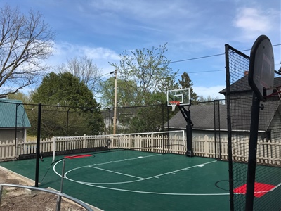 Outdoor sports court with a green surface and red key areas, featuring basketball hoops and a volleyball net, surrounded by a wooden fence.