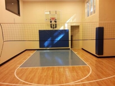 Indoor sports court with a wooden surface and a grey painted area, featuring a basketball hoop and a volleyball net, with wall padding for protection.