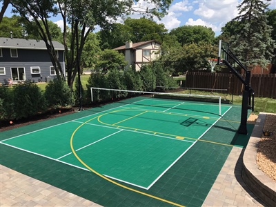 Outdoor multi-sport court with a green surface, featuring a basketball hoop and a volleyball net.