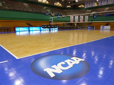 A volleyball court with a wooden playing surface, blue floor around the court with an NCAA logo in Belle Plaine.