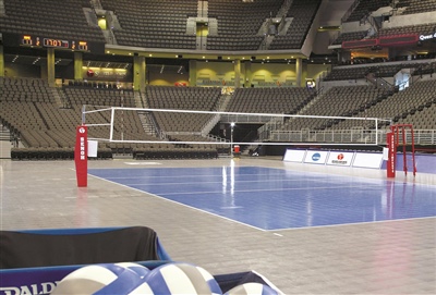 Indoor volleyball court with a blue playing surface and grey surrounding floor. 