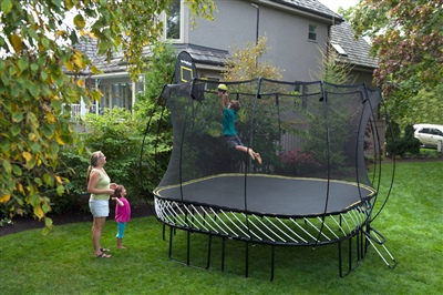 Black trampoline with a black safety net with a yellow spring free layer, a child jumping on it, and a basketball hoop.