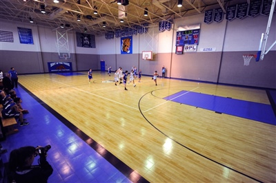 A basketball court with a maple wood-like surface, black lines, and a sport court logo in the center with kids playing basketball.