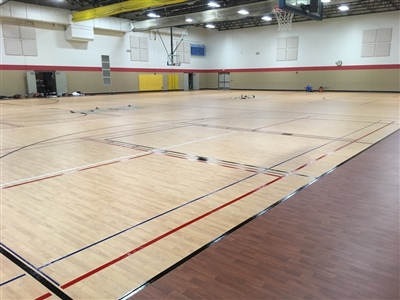 An indoor basketball court with a light maple wood-like surface, featuring multiple colored lines for different sports.