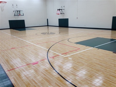 An indoor basketball court with a polished wood surface, green key areas, and a central logo, featuring white and black lines.