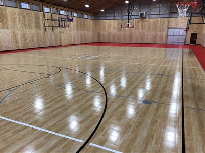 An indoor basketball court with a polished wood surface, black lines, red key areas, and a central logo, surrounded by wooden walls.