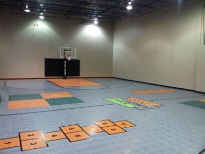  An indoor basketball court with a grey surface, featuring orange and green sections for various sports activities, and a basketball hoop at one end.