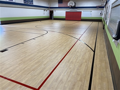 An indoor basketball court with a light wood surface, black and red lines, green and brown walls, and a basketball hoop at one end.