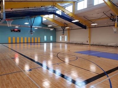 An indoor basketball court with a polished wood surface, blue key areas, black lines, and bright yellow structural beams overhead.