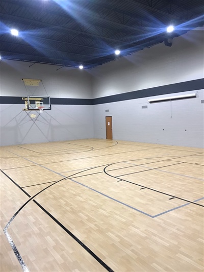 An indoor basketball court with a light wood surface, black lines, and grey walls, featuring a basketball hoop at one end and bright overhead lighting.