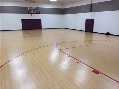 An indoor basketball court with a light wood surface, red lines, and white walls, featuring a basketball hoop and a maroon padded area at one end.