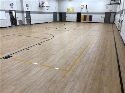 An indoor basketball court with a light wood surface, black and yellow lines, and white walls, featuring multiple basketball hoops.