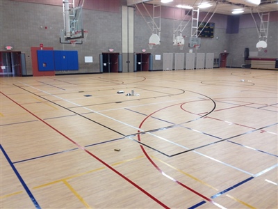 An indoor basketball court with a light wood surface, multi-colored lines, and grey walls, featuring multiple basketball hoops and a central logo in River Hills.