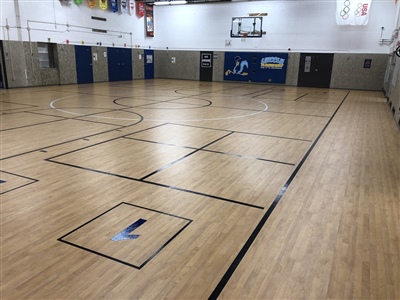 An indoor basketball court with a light wood surface, black lines, and white walls, featuring banners on the walls and multiple basketball hoops.