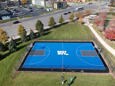 Outdoor residential sports court with a blue and black surface, featuring two basketball hoops and a a Best Buy logo in the center.