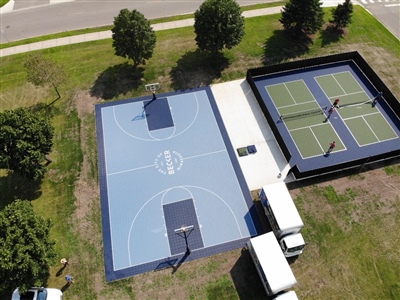 Outdoor residential sports area with a blue basketball court and a separate tennis court, surrounded by grass and trees.