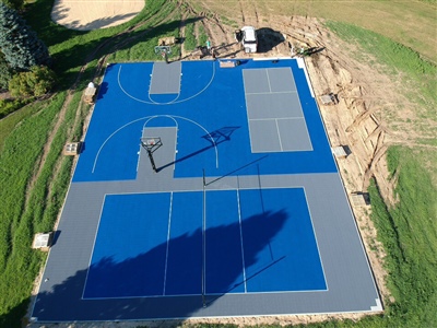 Outdoor residential sports complex with blue and gray surfaces, featuring basketball courts and tennis courts, surrounded by grass and trees.