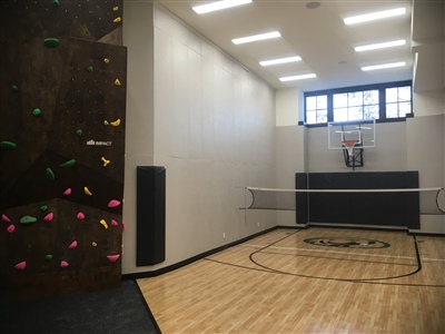  A small indoor basketball court with a light wood surface, black lines, and white walls, featuring a basketball hoop and a rock climbing wall on one side in Mequon.