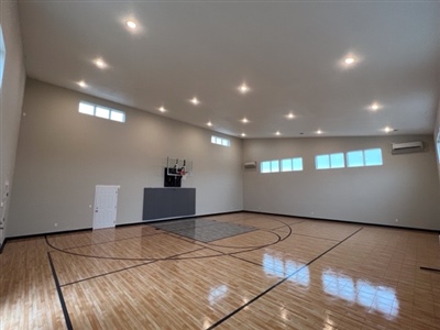 A small indoor basketball court with a polished wood surface, black lines, and light grey walls, featuring a basketball hoop and bright overhead lighting.