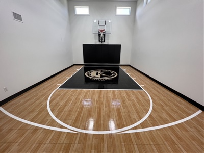 A small indoor basketball court with a polished wood surface, white lines, and white walls, featuring a black key area and a basketball hoop with padding behind it.