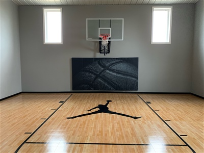 A small indoor basketball court with a polished wood surface, black lines, and light grey walls, featuring a central Jumpman logo on the floor and a basketball hoop with padding behind it.