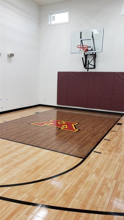 A small indoor basketball court with a polished wood surface, black lines, and white walls, featuring a U of M central emblem on a darker wood section, and a basketball hoop with maroon padding behind it in Maple Bluff.