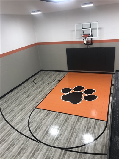 A small indoor basketball court with a grey wood-like surface, black lines, and light grey walls, featuring an orange key area with a black paw print emblem and a basketball hoop with black padding behind it in Brookfield.