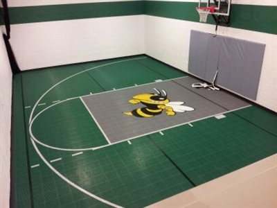 A small indoor basketball court with a green surface, white lines, and white walls, featuring a large bee emblem at the center and a basketball hoop with grey padding behind it in Pewaukee.