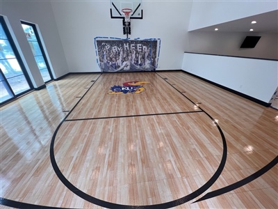  A small indoor basketball court with a polished wood surface, black lines, and white walls, featuring a central logo and a basketball hoop with decorative padding behind it.