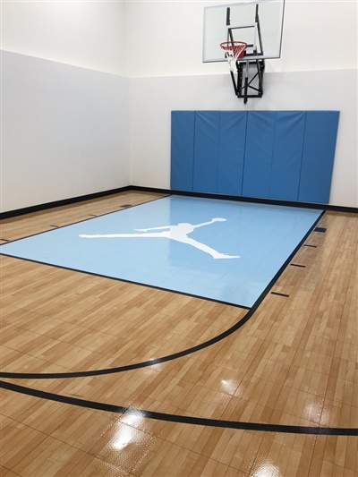  A small indoor basketball court with a polished wood surface, black lines, and white walls, featuring a light blue key area with a white Jumpman logo and a basketball hoop with blue padding behind it in Delafield.