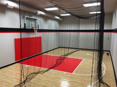 Indoor home basketball court with light wood flooring, red and black wall padding, a basketball hoop, and a black net hanging from the ceiling.