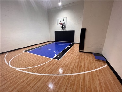 Indoor home basketball court with polished wood flooring, a blue painted key area, black wall padding, and a basketball hoop.