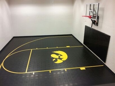 Indoor home basketball court with black flooring, yellow lines, a yellow Iowa Hawkeyes logo, a basketball hoop, and black wall padding.