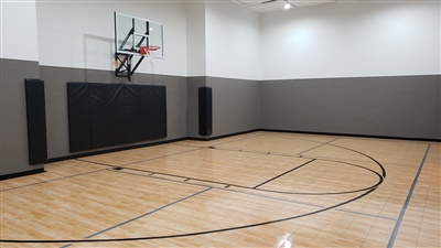 Indoor home basketball court with polished wood flooring, black lines, a basketball hoop, and gray walls with black padding in Middleton. 