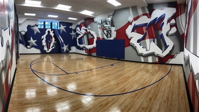 Indoor home basketball court with polished wood flooring, graffiti-style wall art in red, white, and blue, and a basketball hoop.