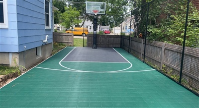 Backyard basketball court with green and gray outdoor basketball flooring, featuring a hoop and netted fence, ideal for residential play and practice.