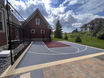 A grey and maroon backyard basketball court with high-quality outdoor basketball flooring, nestled between houses and complemented by a scenic view and clear blue sky.