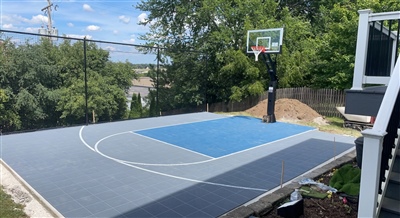 A well-designed backyard basketball court in Lake Elmo with durable outdoor basketball flooring, featuring a blue and grey color scheme, surrounded by trees and a fence for privacy.