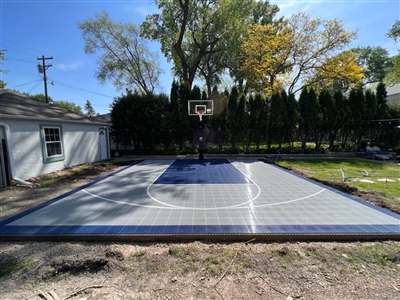 A spacious backyard basketball court in Shorewood, MN featuring high-quality outdoor basketball flooring in grey and navy, set against a backdrop of lush trees and a nearby garage.