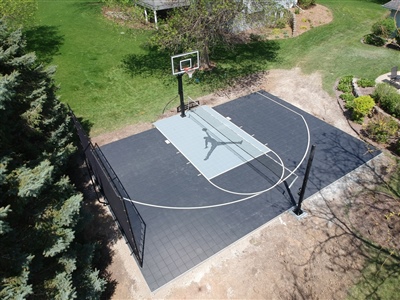 An aerial view of a backyard basketball court in North Oaks, MN with professional-grade outdoor basketball flooring, featuring a central Jumpman logo, set amidst a lush green landscape.