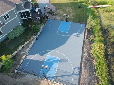 Aerial view of a spacious backyard basketball court in Minnetonka, MN with professional outdoor basketball flooring in grey and blue, adjacent to a well-maintained home and lush greenery.