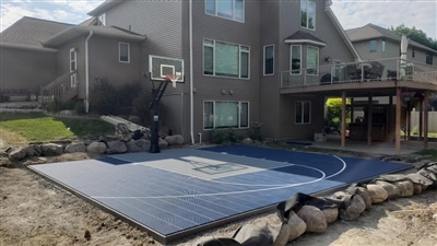 A residential basketball court with a blue and grey surface and white lines surrounded by a rock retaining wall in Green Bay.