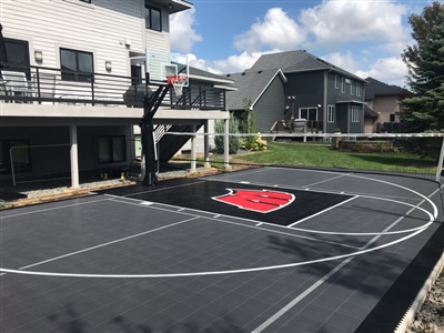 A basketball court with a black and grey surface, white lines, a volleyball net, and a sport court logo in the center.