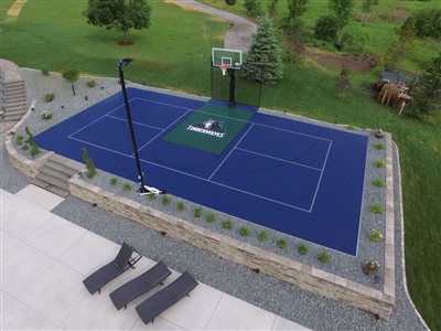  Outdoor residential sports court with a blue and green surface, a basketball hoop, surrounded by a stone retaining wall, and greenery in the background in Madison.