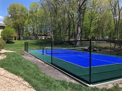 Outdoor residential sports court with a blue and green surface, a black fence, and a tennis net, surrounded by trees and a grassy yard.